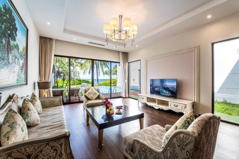 Phòng khách với bộ sofa, bàn trà, tivi và cửa kính lớn nhìn ra vườn và biển.