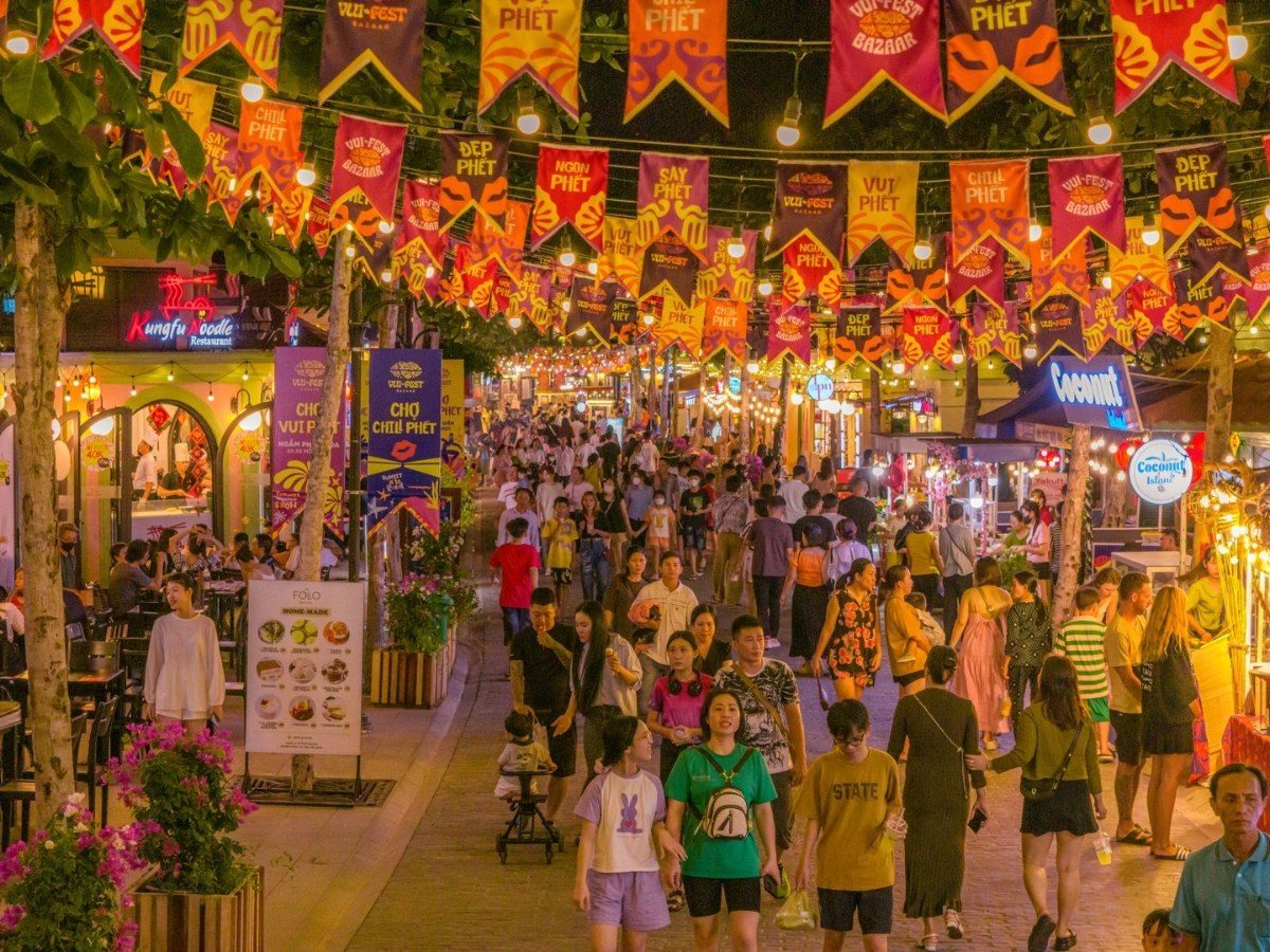 Con phố chợ đêm đông người đi bộ, phía trên giăng nhiều hàng cờ đuôi nheo màu cam đỏ in chữ Vui Phết, hai bên là quầy hàng và cây xanh
