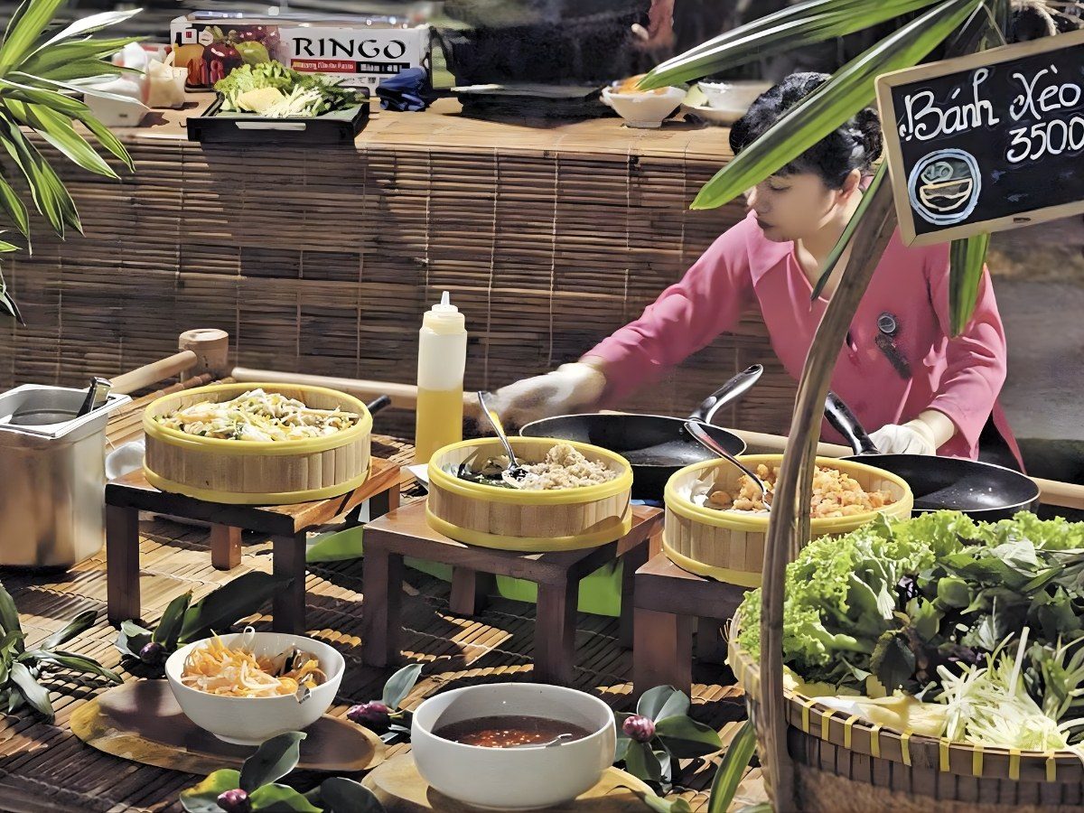 Người bán mặc áo bà ba hồng đổ bánh xèo trên chảo, phía trước là rổ rau sống, chén nước chấm và tấm bảng đen ghi Bánh xèo
