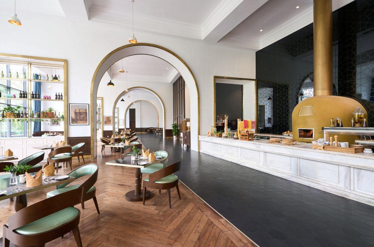 Nhà hàng có vòm cửa trắng, bàn ghế xanh, sàn gỗ, quầy buffet dài và lò nướng màu vàng.
