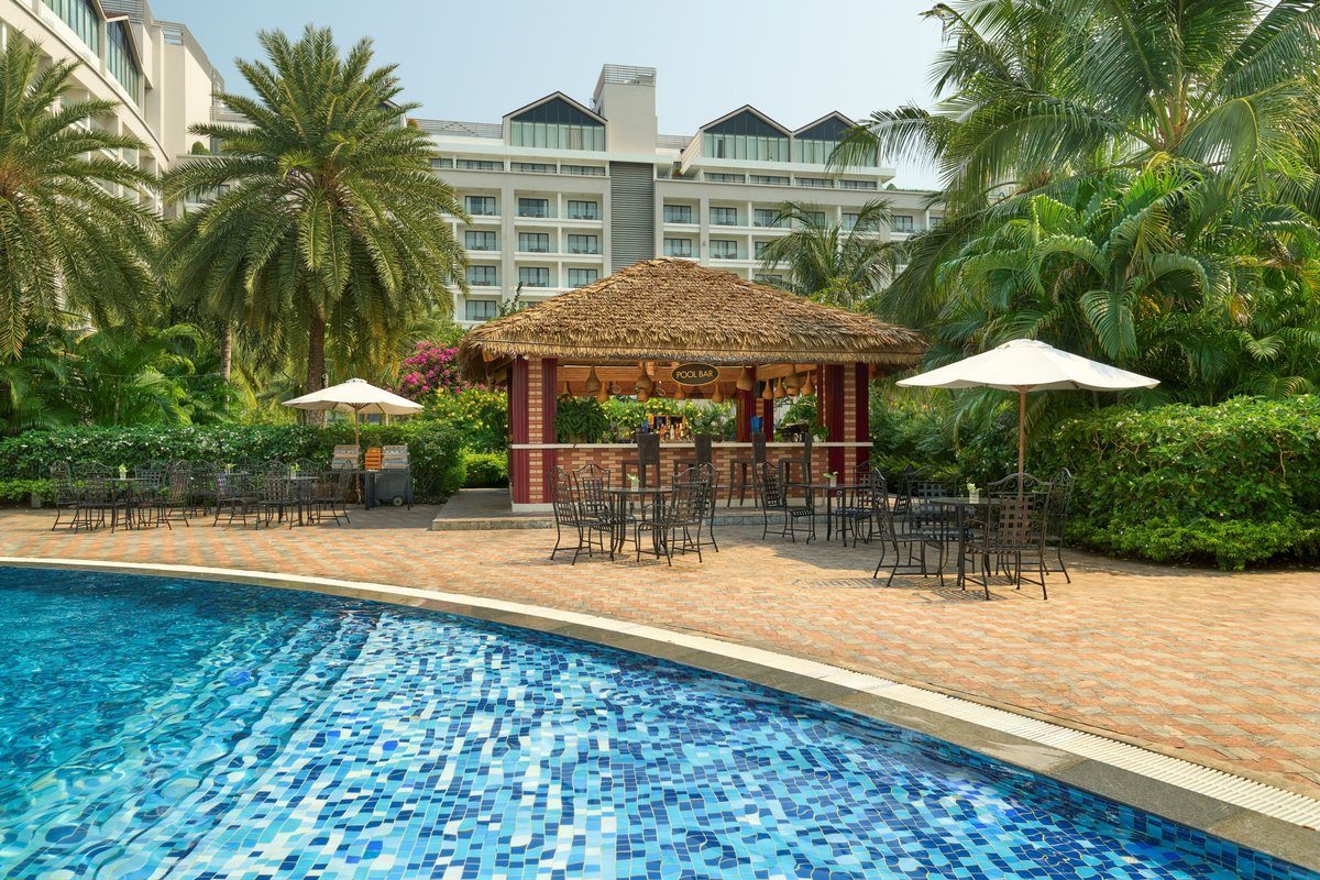 Quầy bar mái lá cạnh hồ bơi lát gạch xanh, có bàn ghế và dù trắng, phía sau là tòa nhà khách sạn.